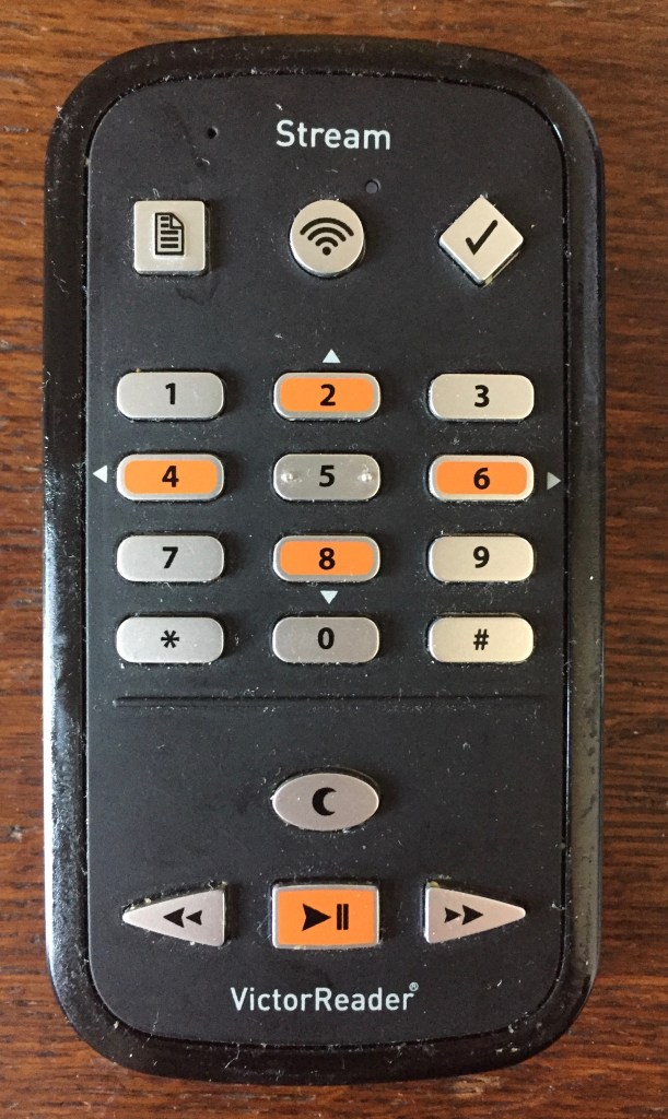 Victor Reader Stream player, a black rectangular device with curved corners. The centre of the device is a number keypad, with 2 dots on the 5. Most keys are silver, but 2, 4, 6 and 8 are orange. At the top are square, circular and diamond shaped buttons. At the bottom is a square orange play button with triangular rewind and fast forward buttons either side, and just above the play button is a silver oval shaped button.