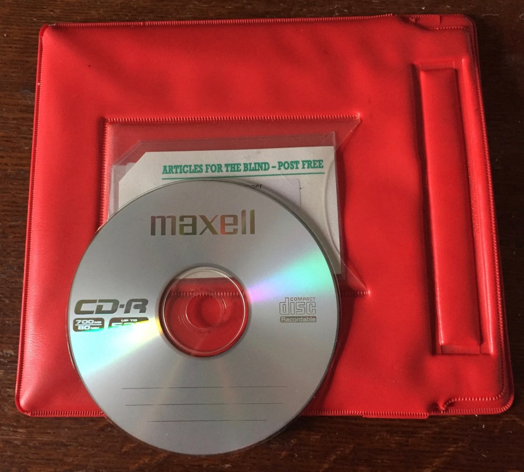 A CD-R disc on top of the red wallet in which it was delivered. The CD is covering the address on the white address card on the wallet, but the top of the card says Articles For The Blind - Post Free.