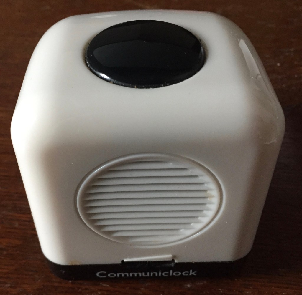 White cube device with curved corners. The front face has a big round speaker, above a small horizontal volume dial, and at the bottom is the word Communiclock. On the top of the cube is a big round black button.