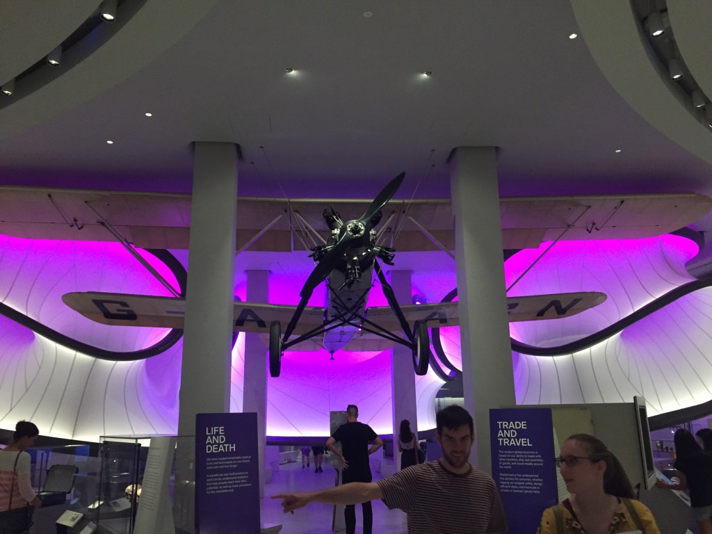 Looking towards the central area of the Mathematics gallery, with curving walls in the background bathed in purple light. Suspended from the ceiling is a large biplane, and below it people are looking at the plane and other exhibits nearby.