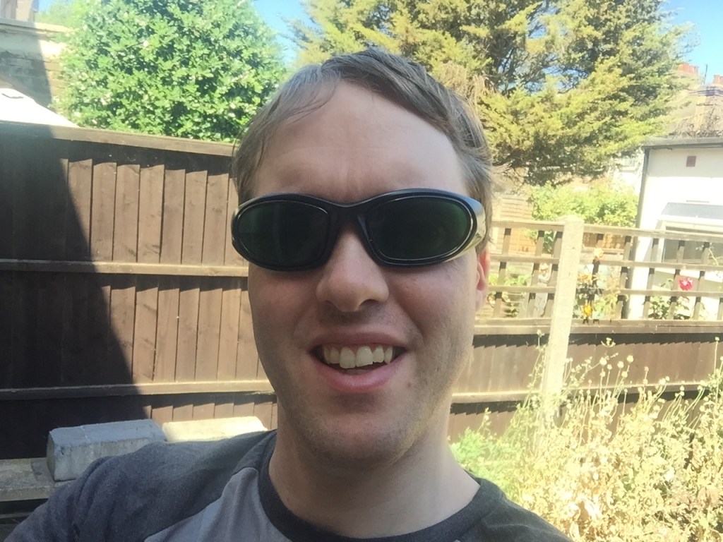 Selfie in my sunny back garden, in which I'm smiling and wearing my green tinted sunglasses.