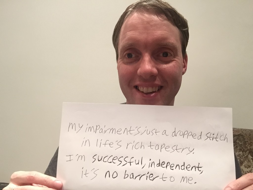 Me smiling and holding up a piece of paper showing a handwritten quote I've come up with. It says: “My impairment’s just a dropped stitch in life’s rich tapestry. I’m successful, independent, it’s no barrier to me.”