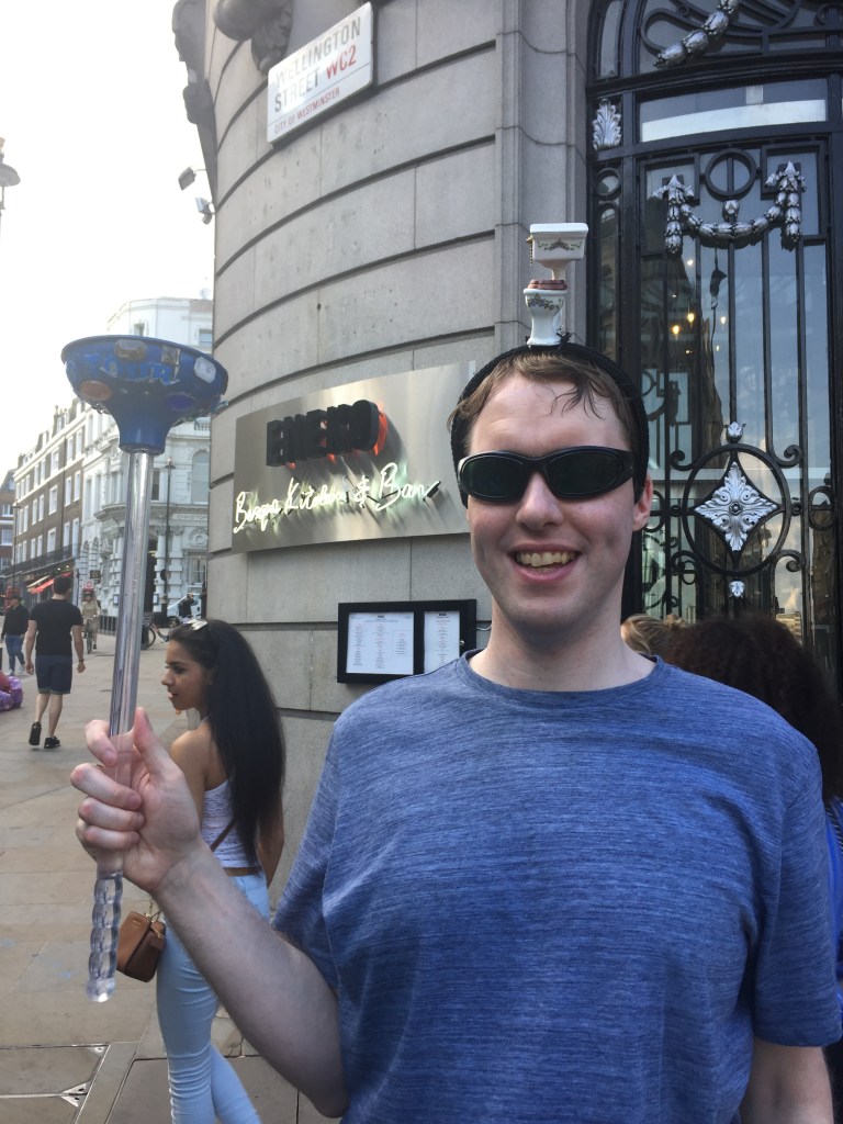 Photo of me outside on a busy London street, smiling and wearing my sunglasses, with a small model toilet on my head and a large plunger in my hand.