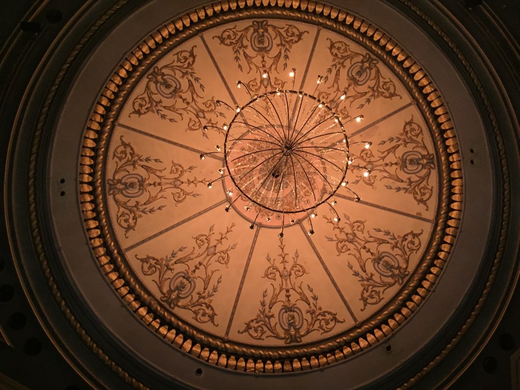 The ceiling of the Old Vic auditorium. The lights in the centre consist of many small bulbs arranged in 2 circles. Around this is a much larger circle on the ceiling itself. divided into 8 equal segments, each containing an ornate leafy design.