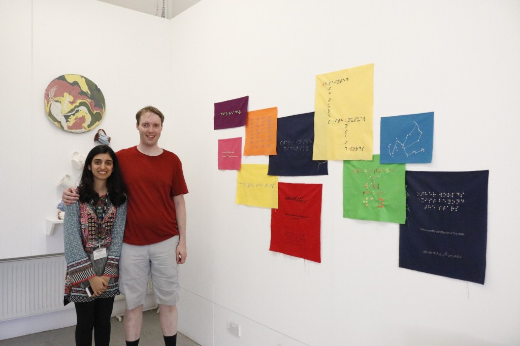 Glen and Nihan standing next to her artwork made up of pieces of coloured fabric on the wall, each a different colour and size, and either square or rectangular in shape, and each containing a message in braille.