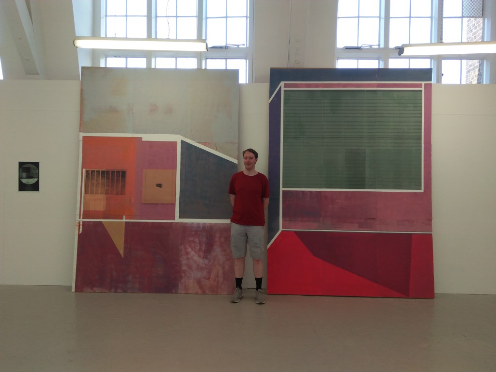 Me standing in front of a very large artwork, considering of large colourful panels, mainly squares and rectangles, but some have diagonal edges on one side.