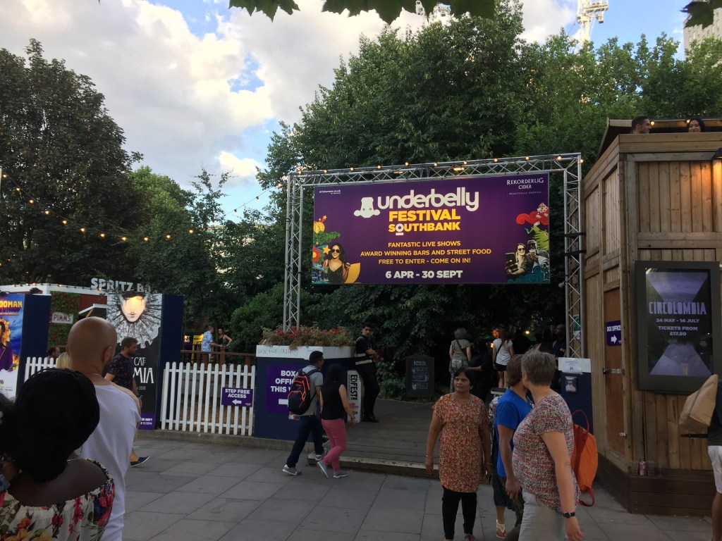 Entrance to the Underbelly Festival on London's South Bank. On the blue sign above the entrance, white and yellow text reads Underbelly Festival Southbank, fantastic live shows, award winning bars and street food, free to enter - come on in, 6 April to 30 September.