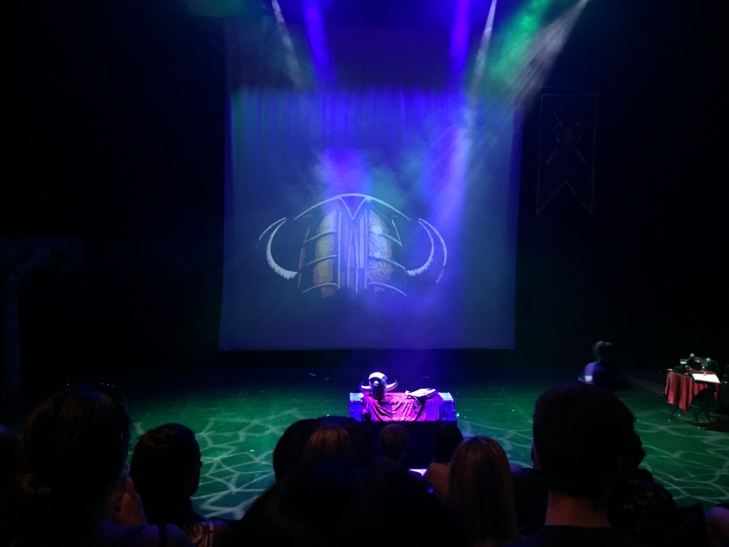 Stage for Knightmare Live. Surrounded in darkness, the large back screen is filled with a large image of the dungeoneer's helmet, which goes down far enough in the front to cover the eyes, and has 2 horns curving inwards on each side. On a table on the stage is an actual dungeoneer's helmet plus a knapsack for carrying items.