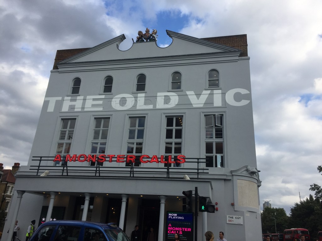 The outside of the Old Vic theatre building, with the words The Old Vic in big white letters across the wall between the rows of windows. Above the theatre entrance are red letters that say A Monster Calls.