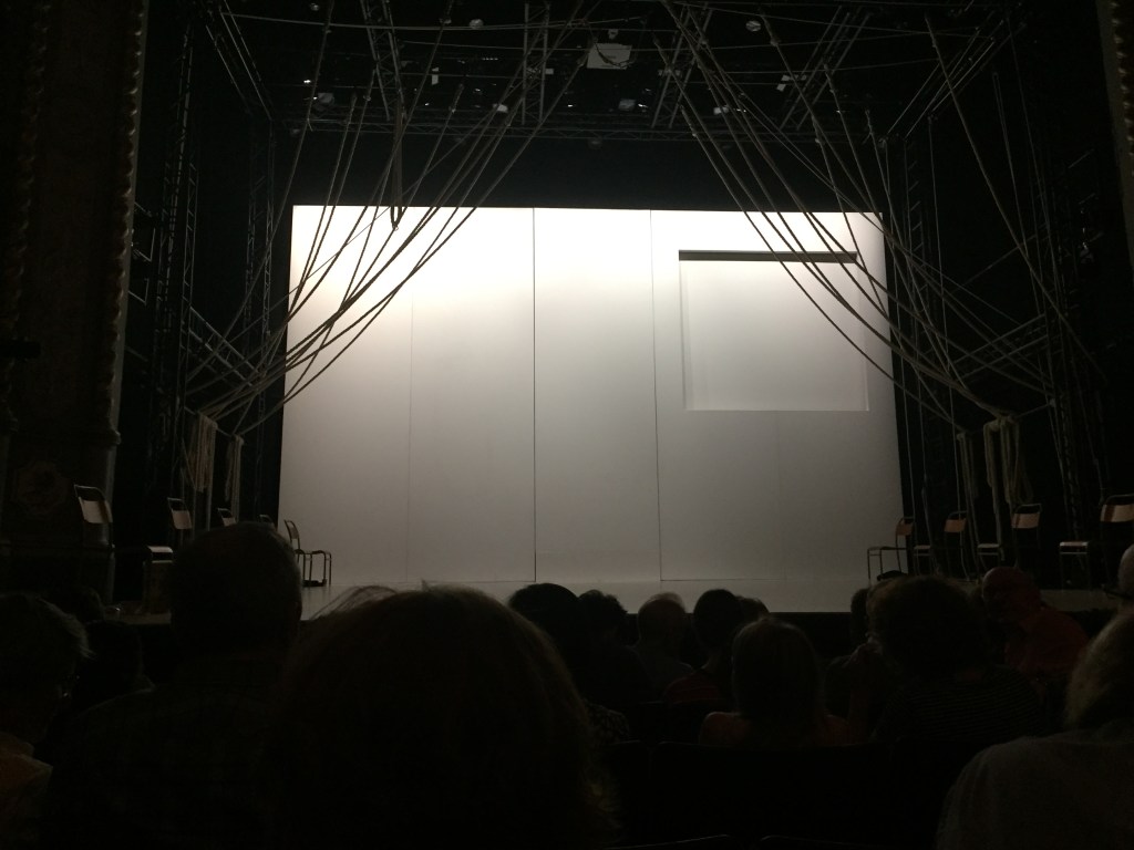 The empty stage for A Monster Calls. The floor and back wall of the stage are white, and at the top right of the back wall is a large square panel inset from the rest of the wall. Lengths of rope extend down from the ceiling and curve across to each side of the stage where they're held in place.