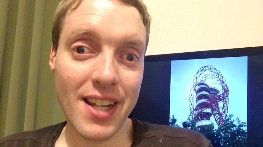 Glen sitting and smiling by his computer, which has a photo of the Arcelormittal Orbit Tower on the screen.