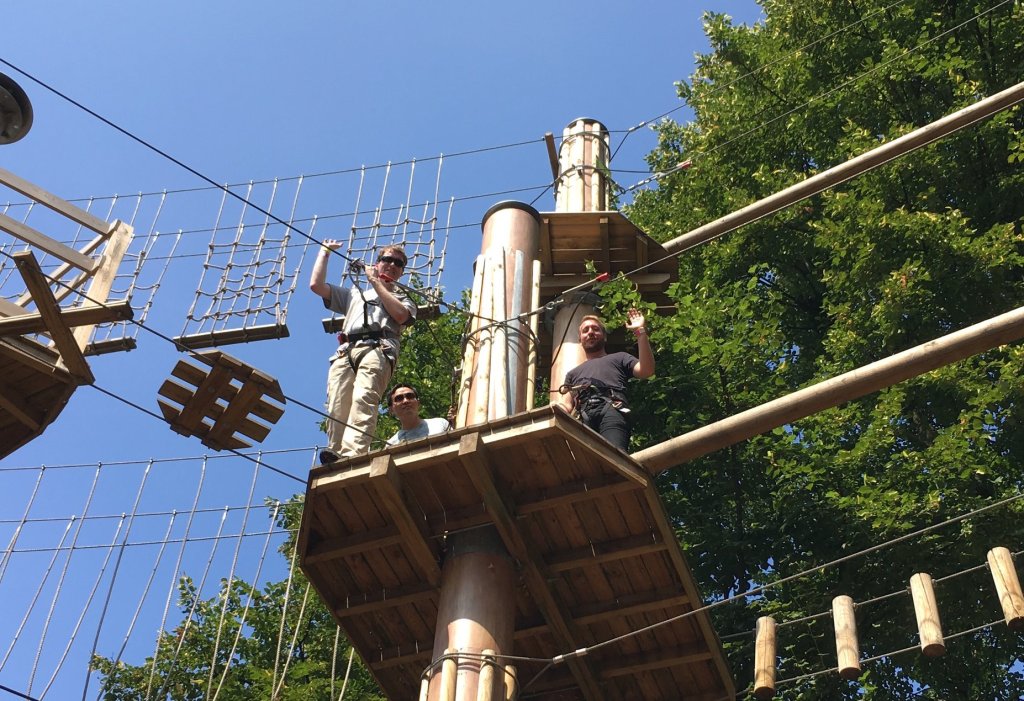 Glen smiling and waving down from a high platform, before crossing a rope bridge that has alternate circular and X shaped platforms. Paul, an East London Vision volunteer, is also waving next to Glen on the platform. Behind them are green trees under the clear blue sky.