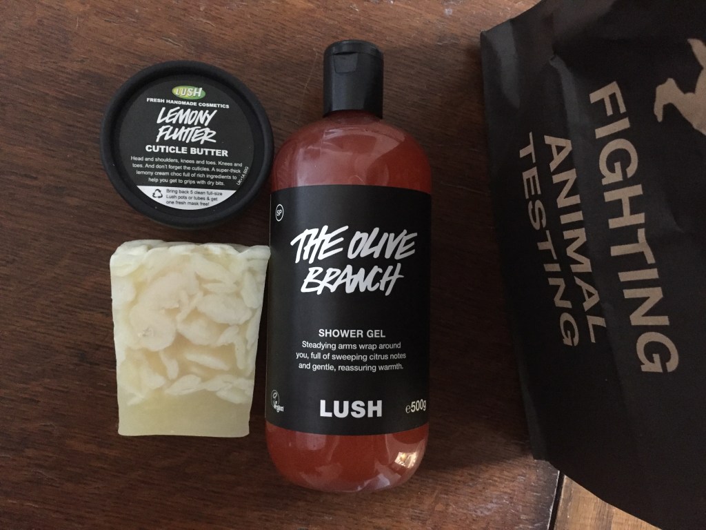 Lush Purchases - a big bottle of The Olive Branch shower gel, the contents of which is red in colour, a bar of the creamy yellow coloured Bohoemian Soap, and a small round pot of Lemony Flutter Cuticle Butter. The items are sat next to a bag that has Fighting Animal Testing on it in capital letters.