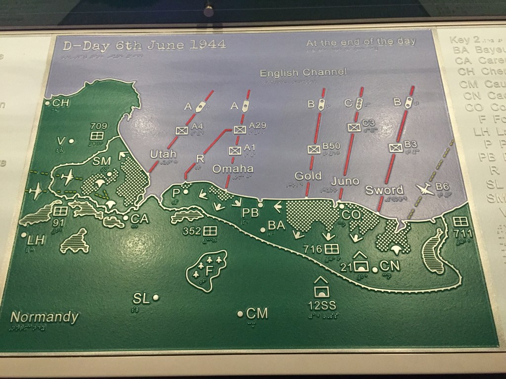 A map of Normandy on D-Day, entitled At The End Of The Day. The land is coloured green with white edging and the sea is in blue. The map is covered in lines and arrows showing the movements of ships in the English channel and personnel on the land. The names of the 5 beaches are also shown in white - Utah, Omaha, Gold, Juno and Sword.
