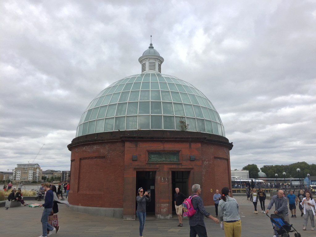 Entrance to the Greenwich Foot Tunnel, a large round brick building with a glass dome made of 5 rows of window panes going all around it, and a small domed spire on the top.
