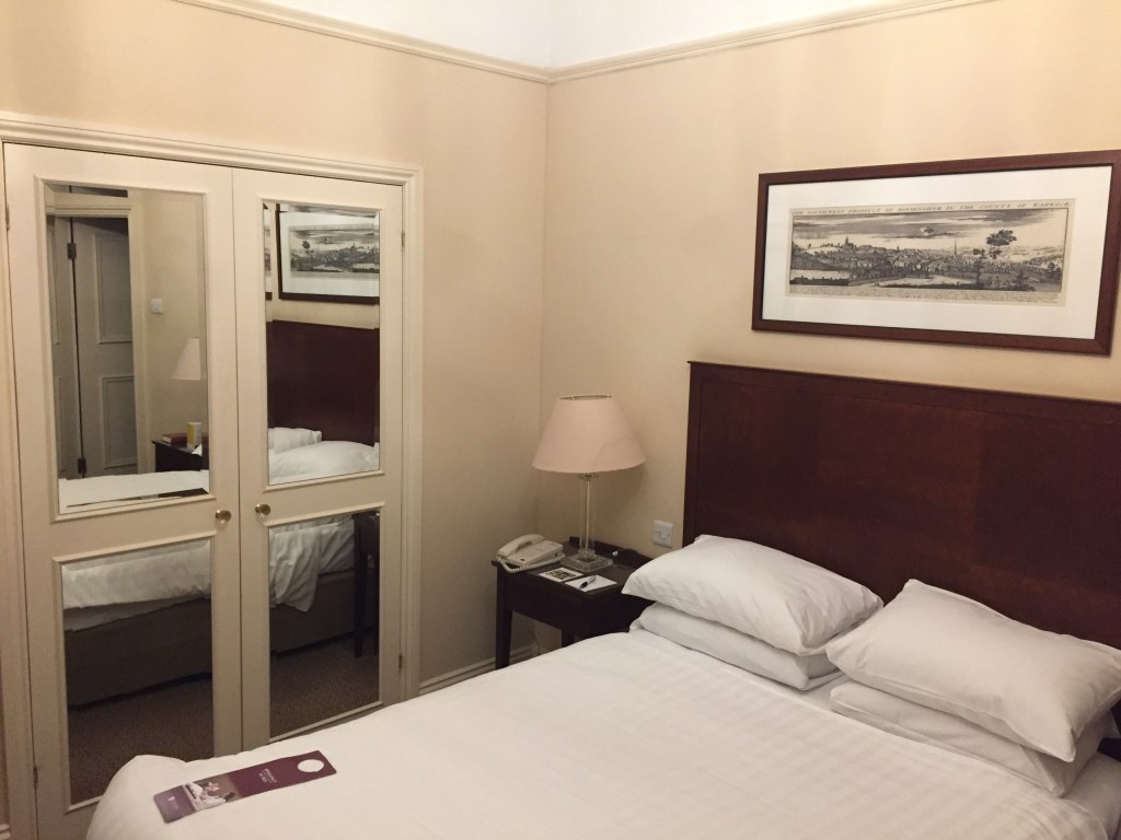 Hotel room with a double-bed next to a wardrobe with mirrored doors. In the reflection on the doors you can see the entrance to the en-suite bathroom on the other side of the room.