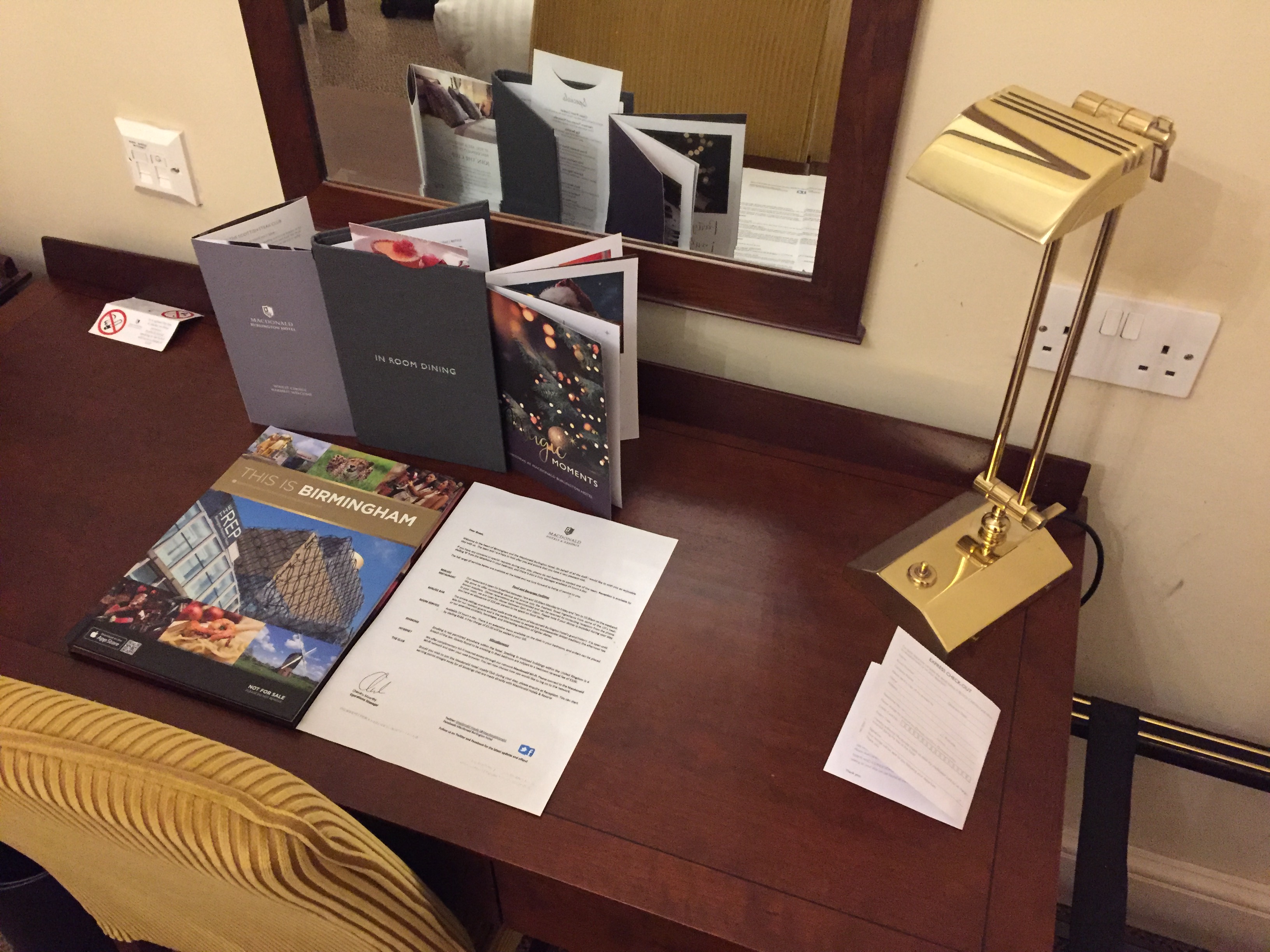 Desk in my hotel rom, with a gold coloured reading lamp, a welcome letter, a hardback book called This Is Birmingham, a leaflet about the hotel, a room service dining menu, and a booklet about their Christmas events. A large mirror is on the wall above the desk.