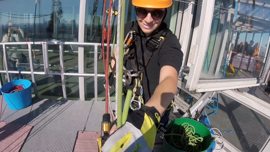 Glen's headcam view of his female abseil instructor. She's smiling as she holds on to the rope in front of her, while guiding him backwards over the edge of the platform. Through the large window behind the instructor we can also see a lady from Moorfields Eye Charity taking a photo of Glen with her phone.