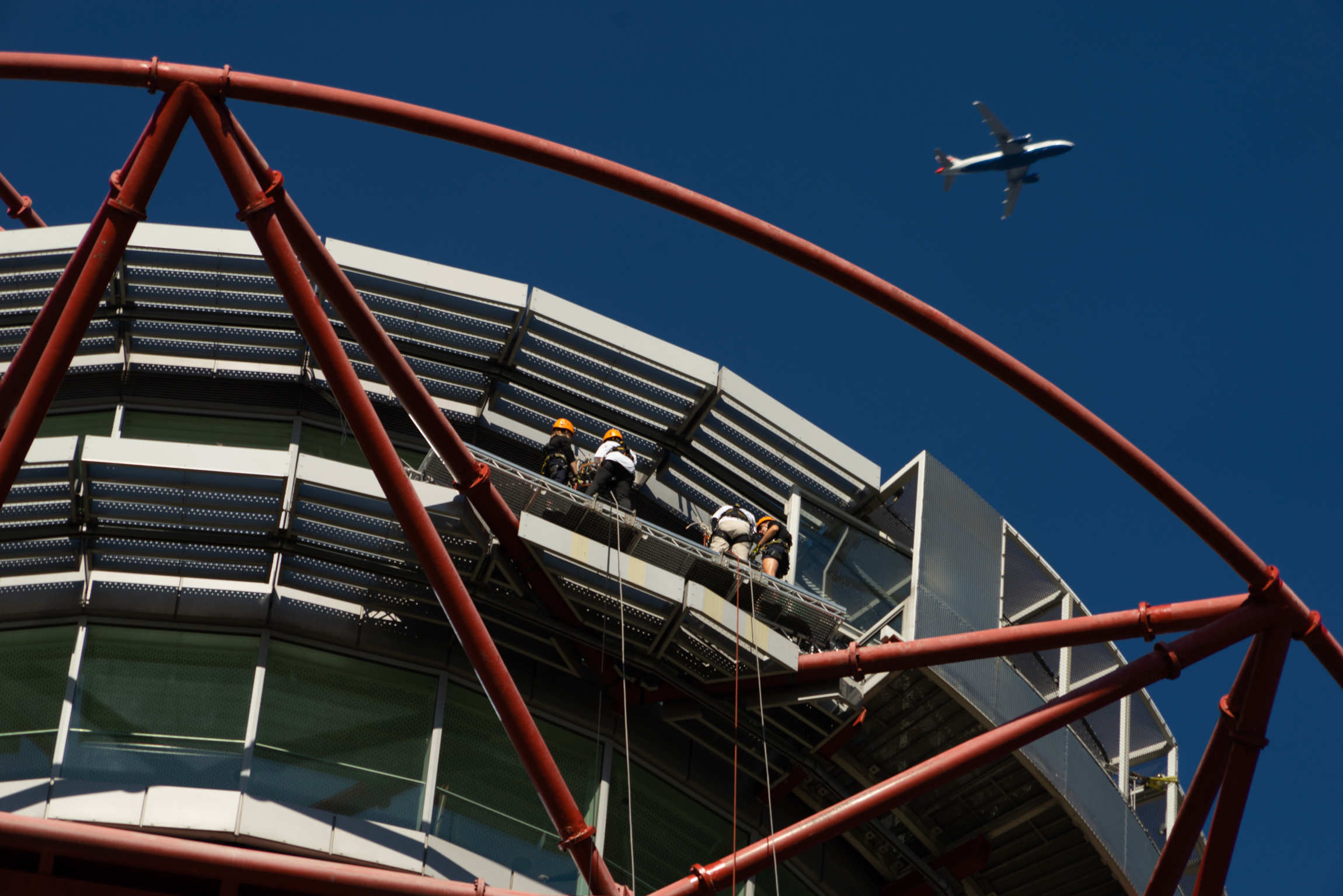 Photo taken by a spectator on the ground, zoomed in on Glen and Claire standing on the abseil platform at the top of the tower, while a large plane flies overhead.