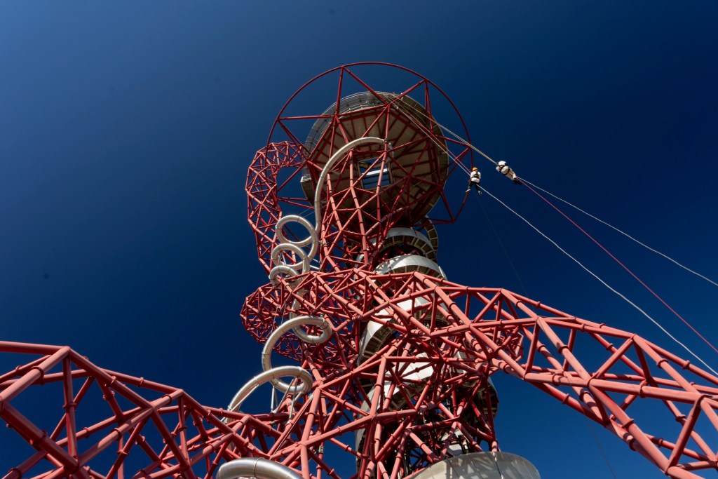 Photo by a spectator on the ground almost directly below the tower, looking up towards the underside of the circular platform at the top. The photo is zoomed out so that most of the tower is visible. Claire and Glen can be seen abseiling on the right side of the tower.