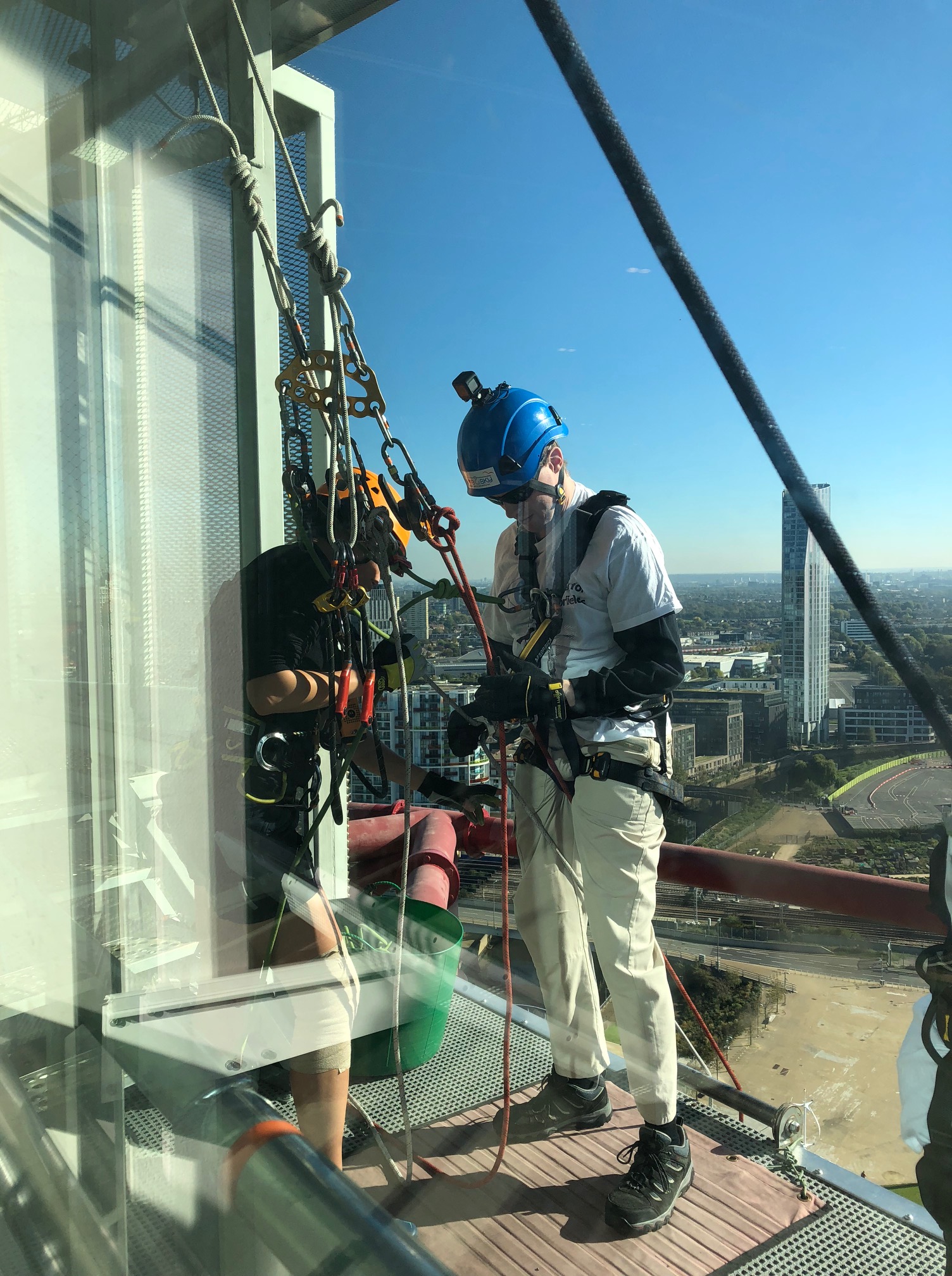 Glen on the abseil platform, with his back to the edge, as the female instructor attaches his harness to the ropes.