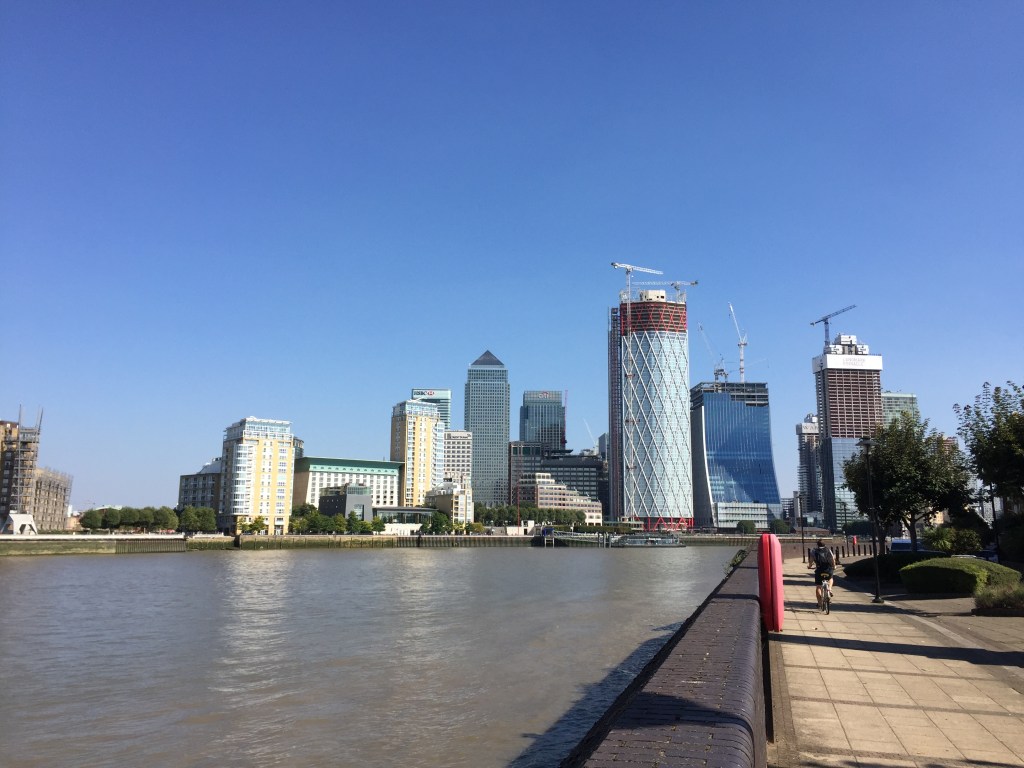 View along the River Thames from the Thames Path. Canary Wharf and other skyscrapers, some with cranes on top, are on the bending curve of the river ahead, under a clear blue sky.