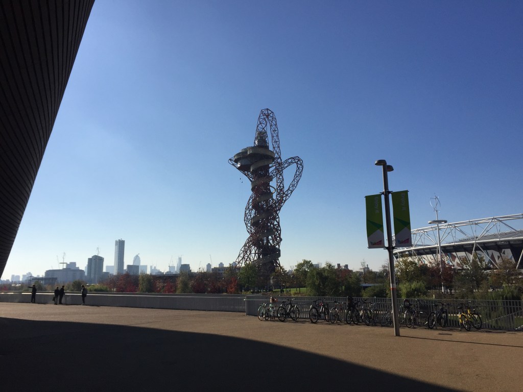 The ArcelorMittal Orbit Tower, a tall, red structure with curving girders that swirl around it, and a curving helter skelter slide from the top to the bottom. Viewed from a distance in this photo, beneath the clear blue sky, you can just see the very tiny silhouettes of 2 abseilers dangling off the left side.