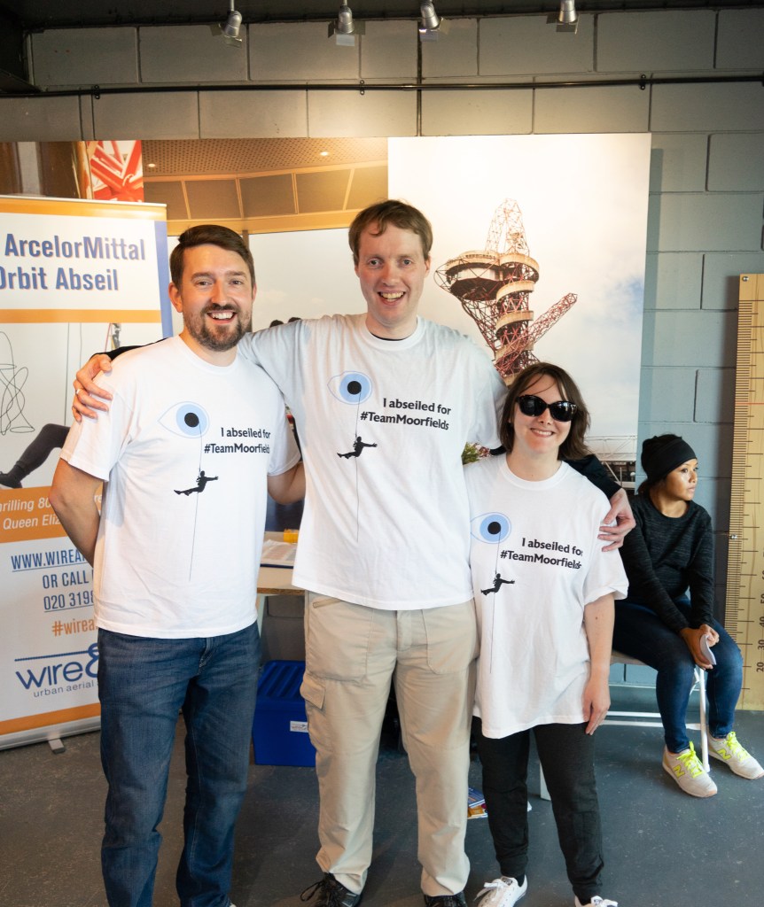 Matt, Glen and Claire posing together and smiling, in their white Moorfields Eye Charity abseil t-shirts. In the centre of the t-shirt, large black text says I Abseiled For hashtag TeamMoorfields. To the left of that is the Moorfields Eye Charity logo - a blue circle with a smaller black circle in the centre, resembling an eye with a pupil. A silhouette of a person is abseiling down a rope from the pupil.