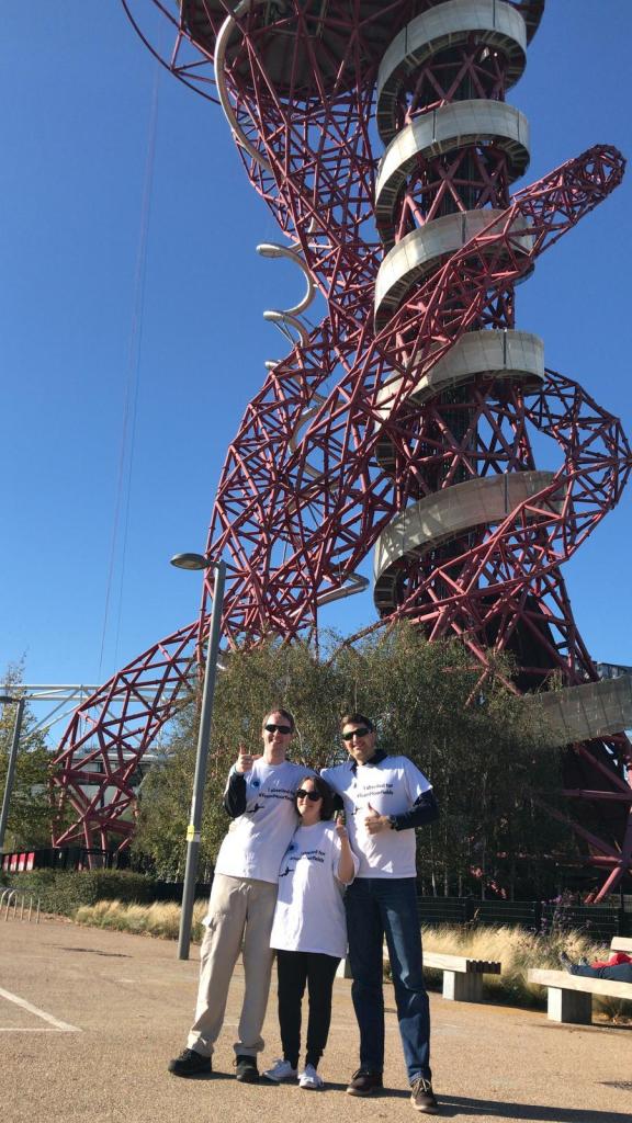 Glen, Claire and James smiling and giving a thumbs up, posing in their Moorfields Eye Charity abseil t-shirts, as the Orbit Tower stands high over them in the background.