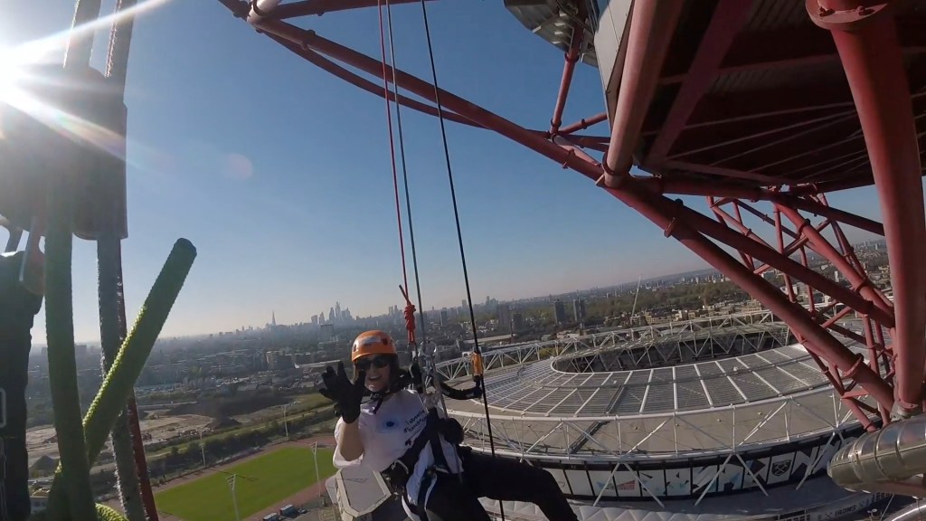 Headcam view to the right of the tower, showing fellow abseiler Claire smiling and waving, with the large round Olympic Stadium behind her, and the London skyline stretching into the distance.