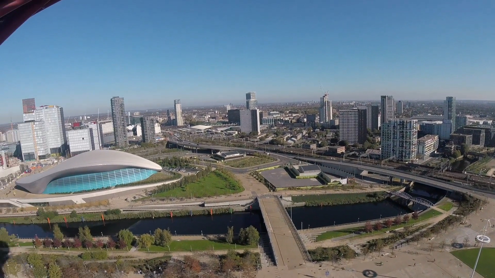Headcam view to the left of the tower. A river flows across the image passing under a couple of pedestrian bridges, a road bridge and a railway bridge, before disappearing between the skyscrapers on the right. On the opposite side of the river to the left is the Aquatics Centre, which isn't very tall, but has a flowing, curving light grey roof along its length, and big blue glass windows along the side.