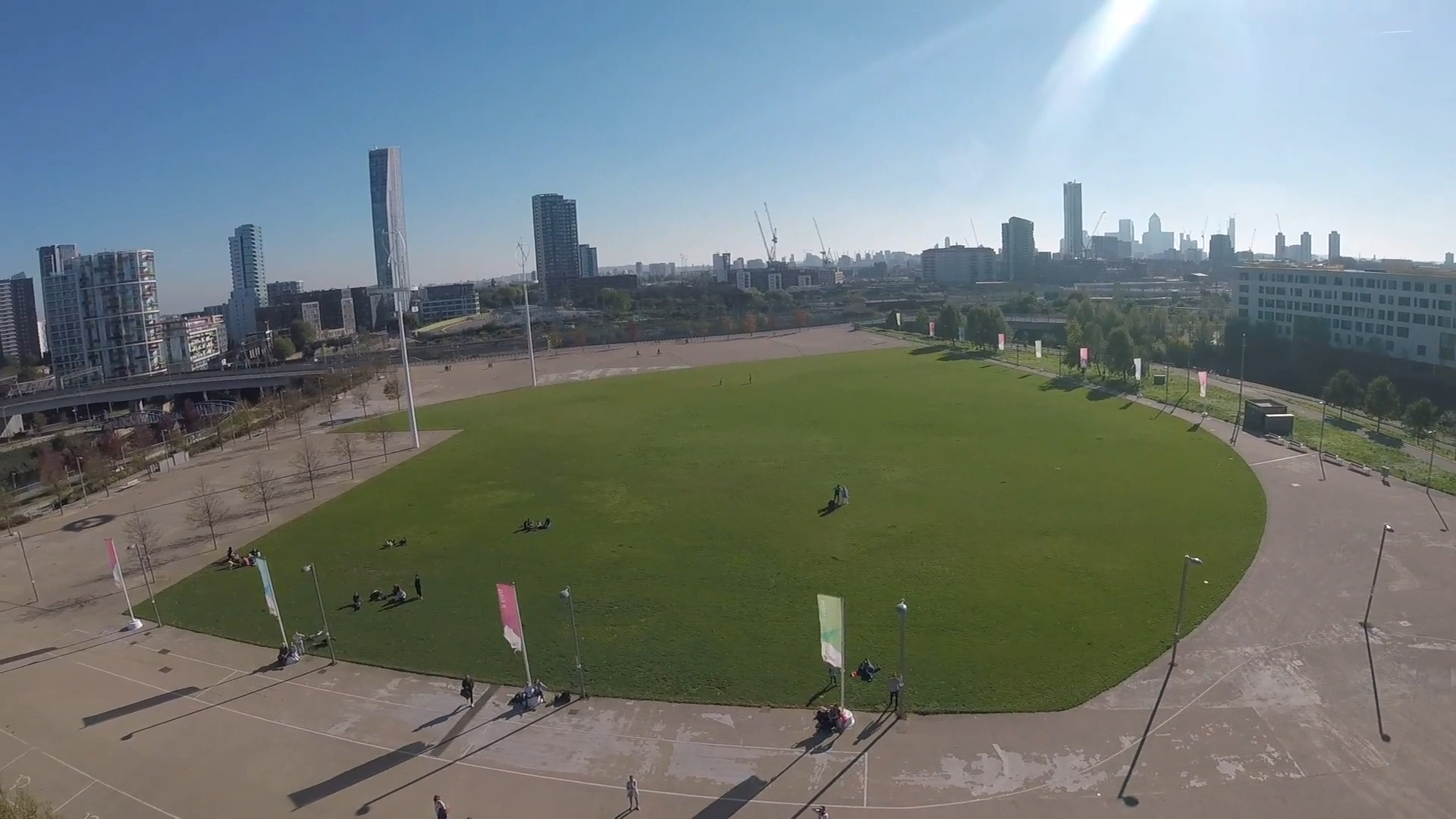 Headcam view of the large round grassy area where spectators can watch the abseil, with a few small trees along the left and right sides, and buildings of various heights across the background in the distance.