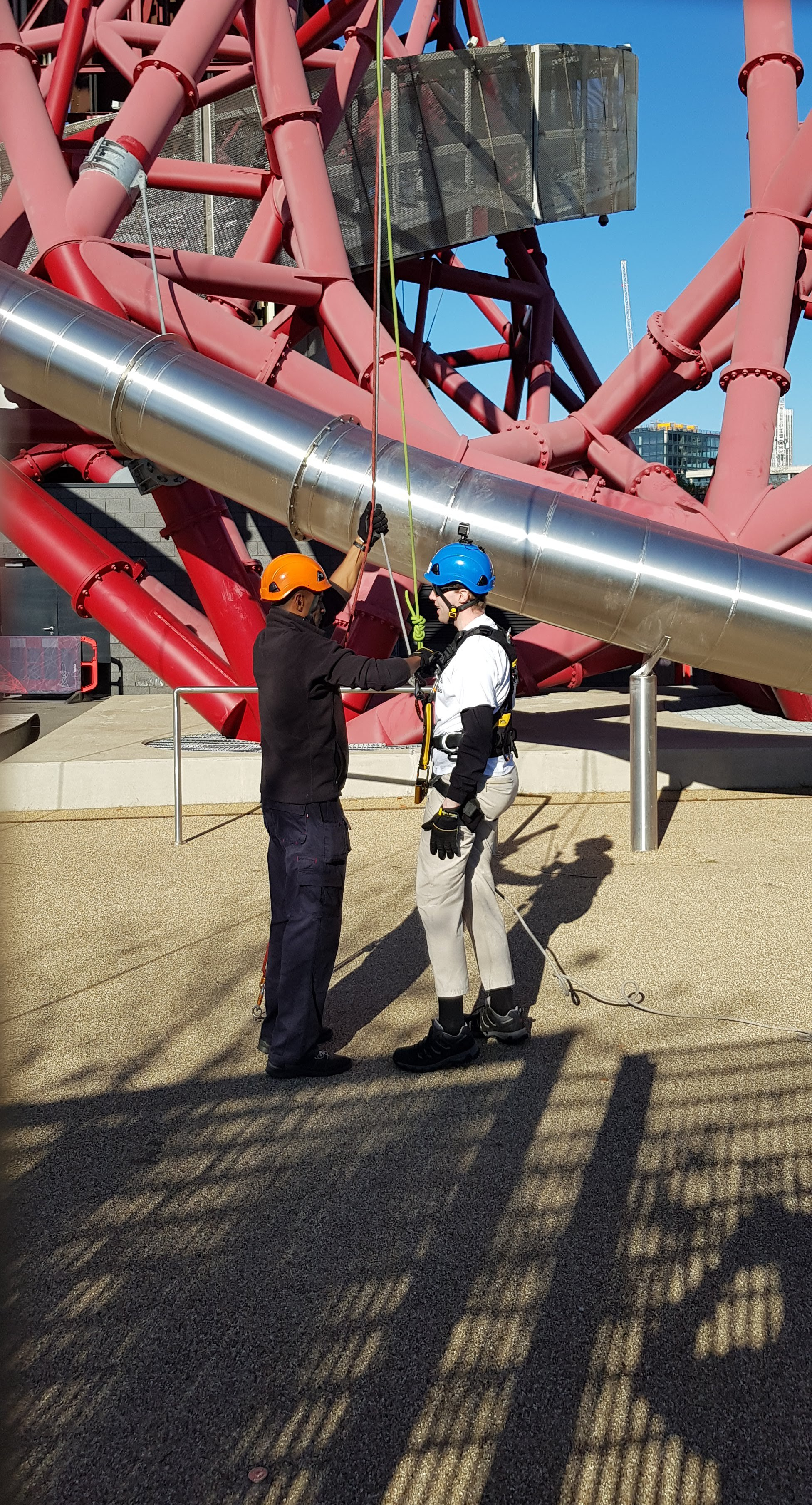 Now standing back on the ground, Glen waits as the male instructor disconnects the ropes from his harness. Part of the large metal slide around the tower can be seen curving towards the ground behind them, the end being somewhere to their right.