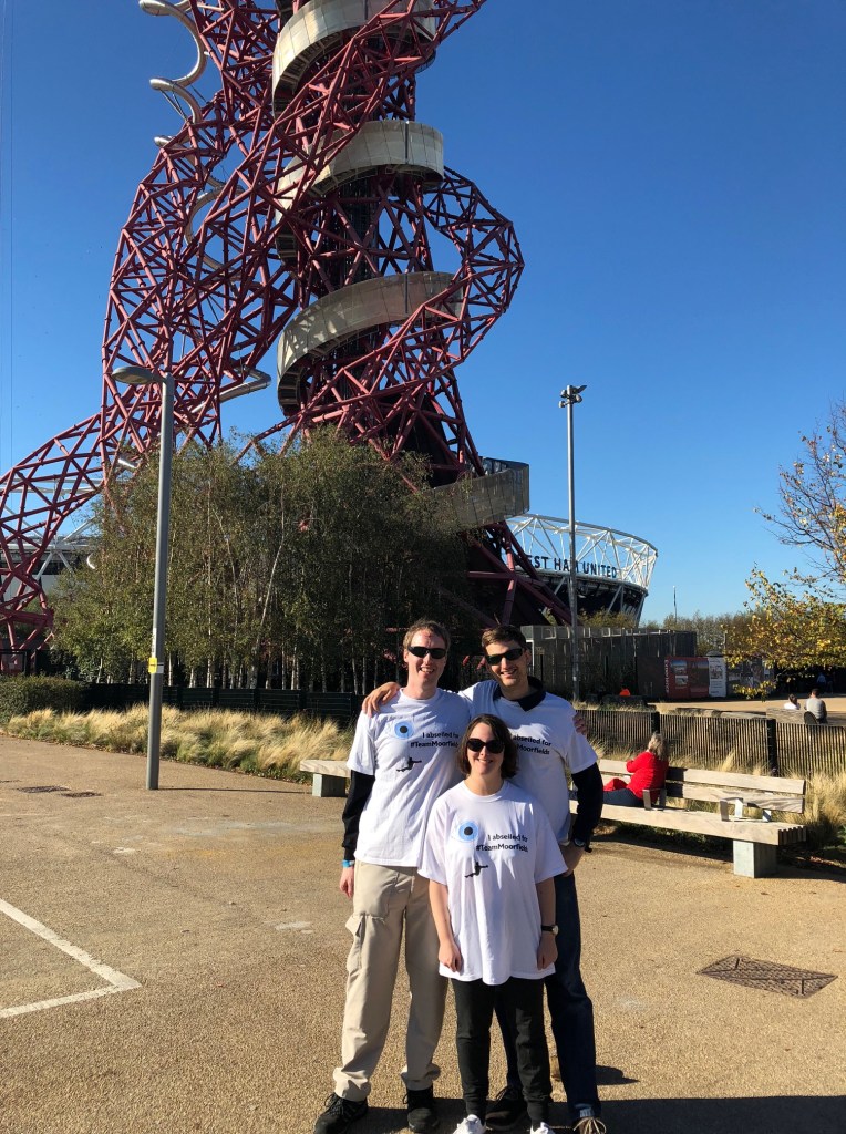 Glen, Claire and James smiling as they pose in their Moorfields Eye Charity abseil t-shirts, in front of the tall Orbit Tower, with the London Stadium visible just behind it.