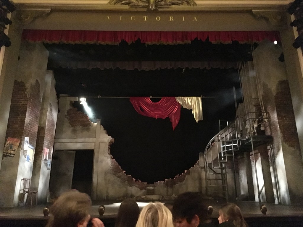 The stage for Much Ado About Nothing, designed to look like a bombed out theatre from the 1940s, with a huge hole in the back wall left behind by a bomb. Torn remnants of green and red fabric hang in the air above it. 2 rectangular pillars on the left of the stage have a few war posters on them, while on the right a spiralling staircase leads up to a balcony.