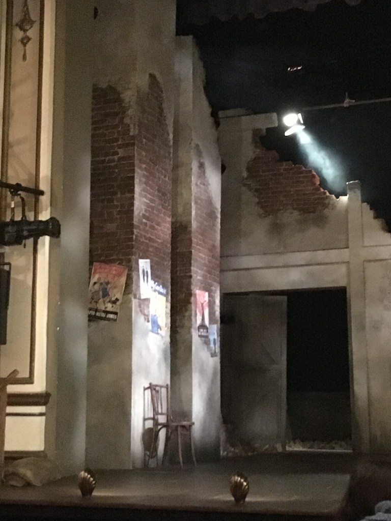 View of the left side of the stage for Much Ado About nothing, showing the 2 tall square pillars. As the set resembles a bombed out theatre from World War 2, the pillars are damaged, with a lot of the white paintwork stripped away revealing the bricks underneath. On the bottom third of the pillar, where most of the white paint remains, various posters from the war are attached.