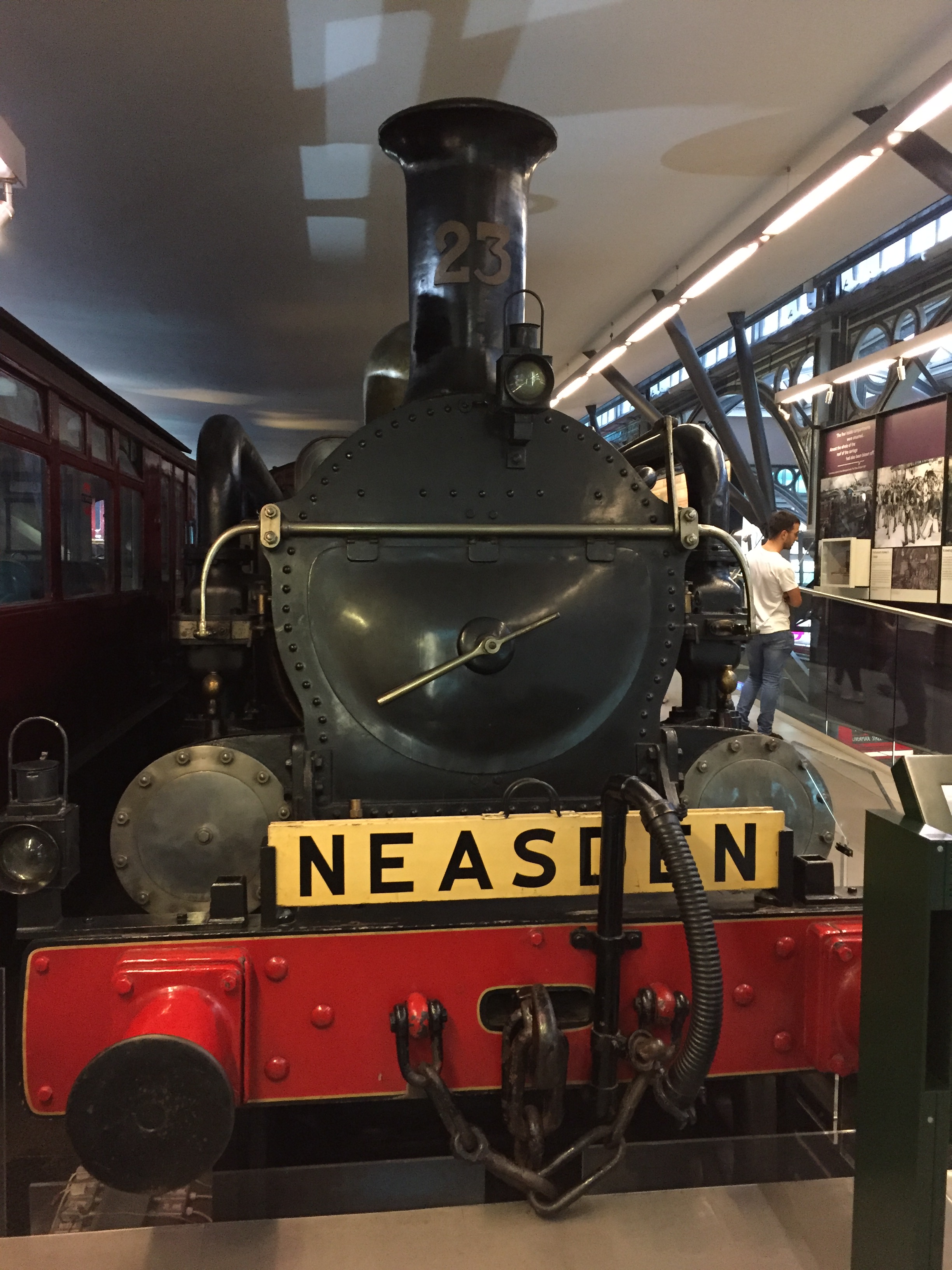 Front view of a Metropolitan Railway Steam Locomotive. The front engine chamber is large, round and black with a tall chimney on top. Below it is a white registration plate with black text saying Neasden. And below that is a red panel with a circular buffer sticking out on each end.