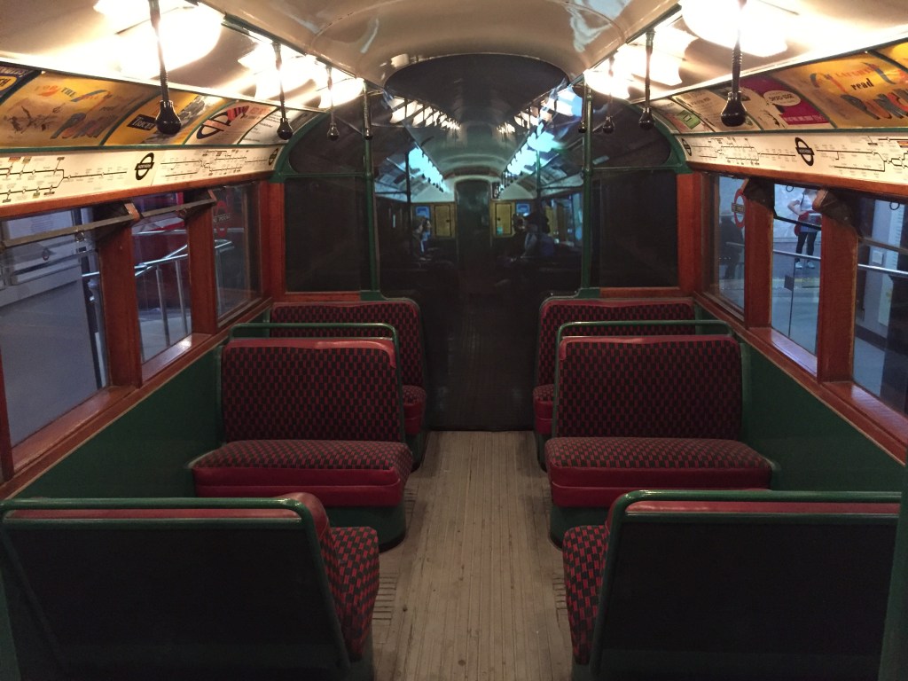 The interior of a 1938 stock tube carriage. The red seats are aligned across the carriage with a small aisle between them, unlike modern Tube trains where seats are aligned along the sides of the carriage. Above the windows on each side of the carriage is a map of the Tube line and various advertising posters.