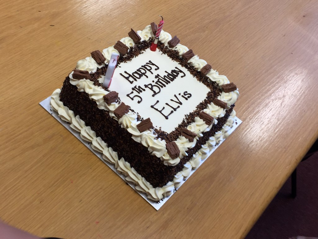 Large square birthday cake, made of chocolate sponge with decorative cream borders around the bottom and top of the cake. The top has white icing on which chocolate lettering says Happy 5th Birthday ELVis.