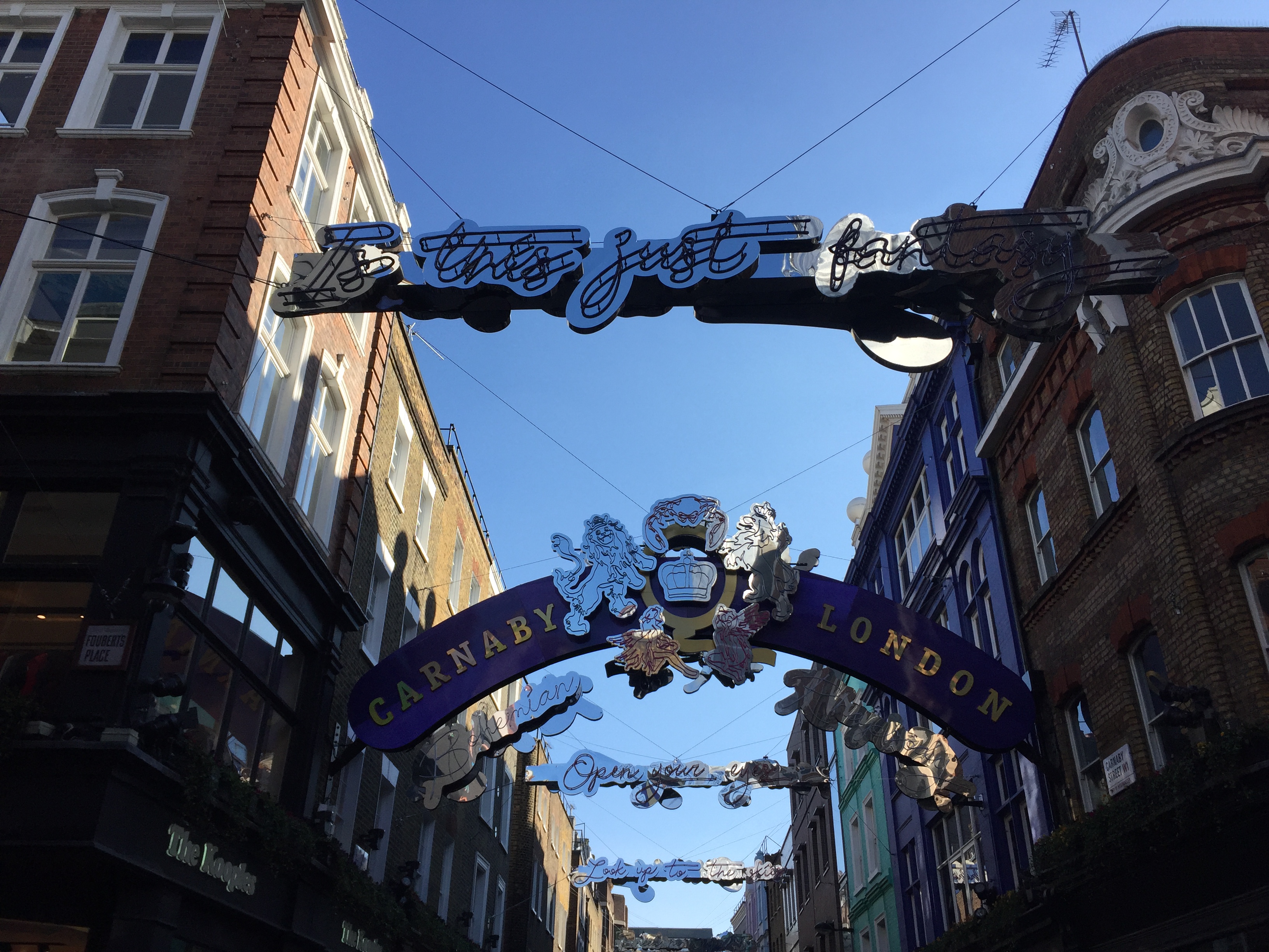 In the daylight, decorations promoting Queen in the air across Carnaby Street. A silver version of the band's crest, with a lion on each side of the central crown, sits in the centre of a curving blue archway that says Carnaby on the left and London on the right in gold letters. Other decorations stretched across the street quote lyrics from Bohemian Rhapsody in curly script lettering, e.g. Is This Just Fantasy, Open Your Eyes, and Look Up To The Skies.