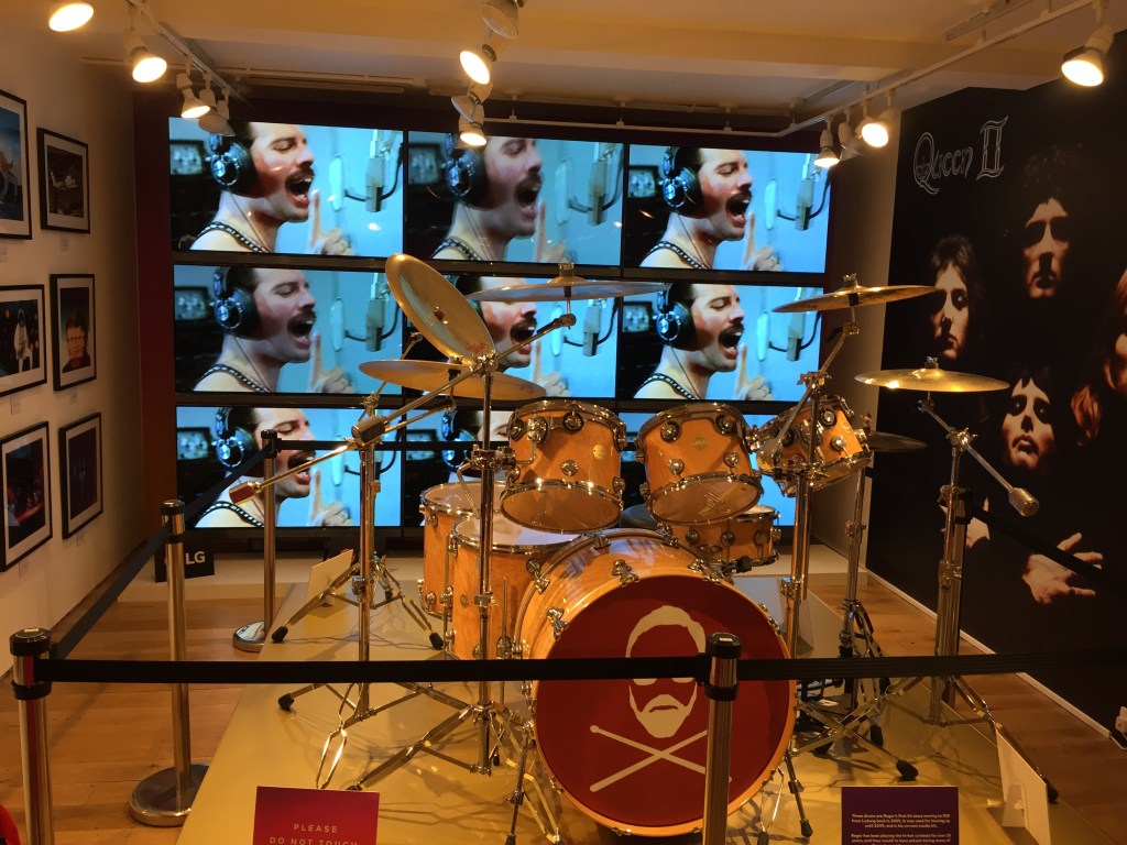 A drum kit used by Roger Taylor on display in the Queen pop-up shop. The front of the drum in the centre has a simple silhouette of Roger Taylor's head among 2 drumsticks laid in an X shape. Behind the drum kit, a grid of 9 screens are all showing the same video of Queen recording One Vision in the studio, with Freddie Mercury singing into the microphone. To the right of the kit and screens is a large poster of the Queen 2 album cover, showing all 4 members of the band.