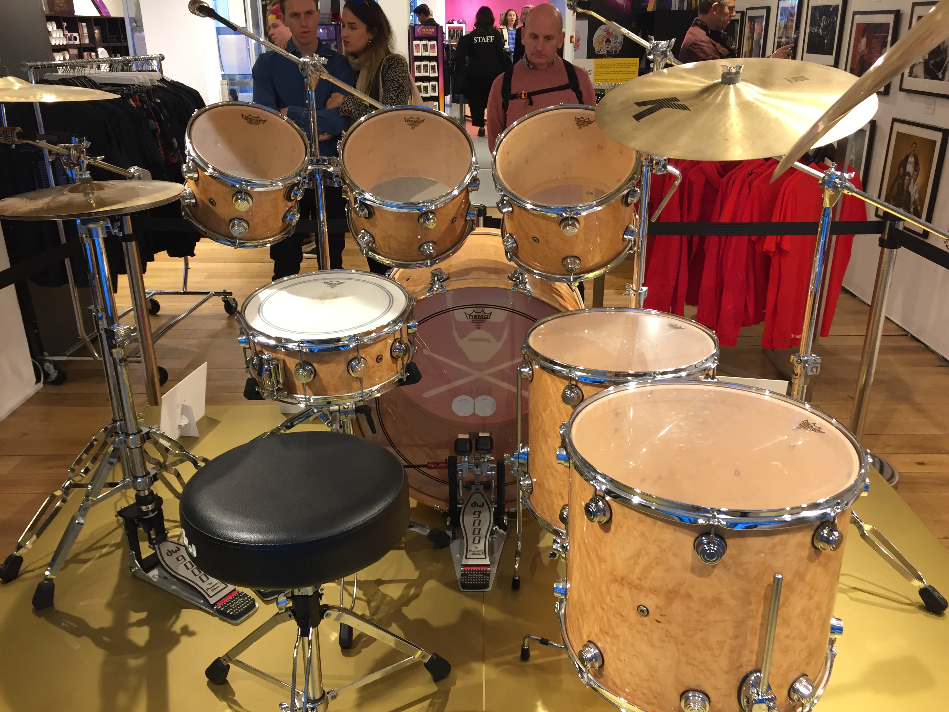 Rear view of Roger Taylor's drum kit in the Queen shop, showing the stool he would sit on and the view of the drums and cymbals that he would have had. There are 2 hi-hat cynbals on the left and 2 larger symbols on the right. Above the bass drum in the centre are 3 drums angled towards the drummer, , and another smaller drum at a flatter angle just in front of the stool. To the right are 2 more large drums below the big cymbals.