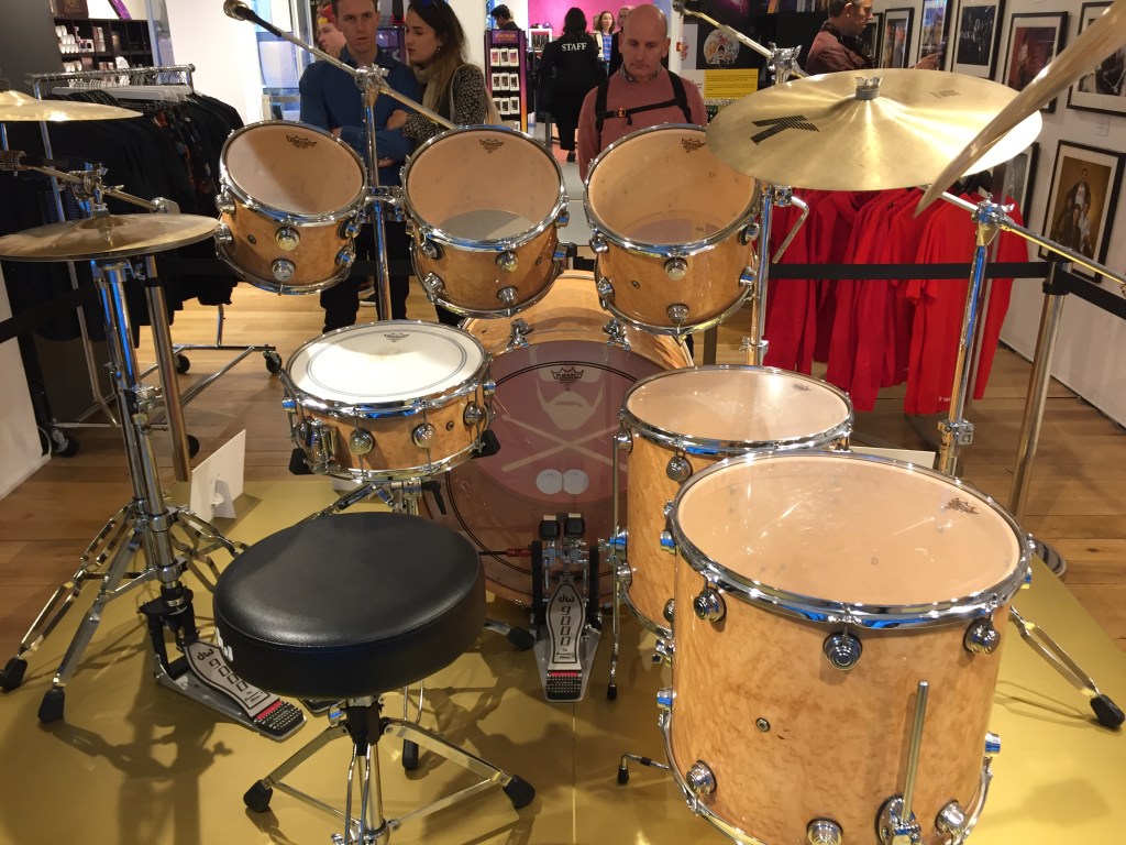Rear view of Roger Taylor's drum kit in the Queen shop, showing the stool he would sit on and the view of the drums and cymbals that he would have had. There are 2 hi-hat cynbals on the left and 2 larger symbols on the right. Above the bass drum in the centre are 3 drums angled towards the drummer, , and another smaller drum at a flatter angle just in front of the stool. To the right are 2 more large drums below the big cymbals.