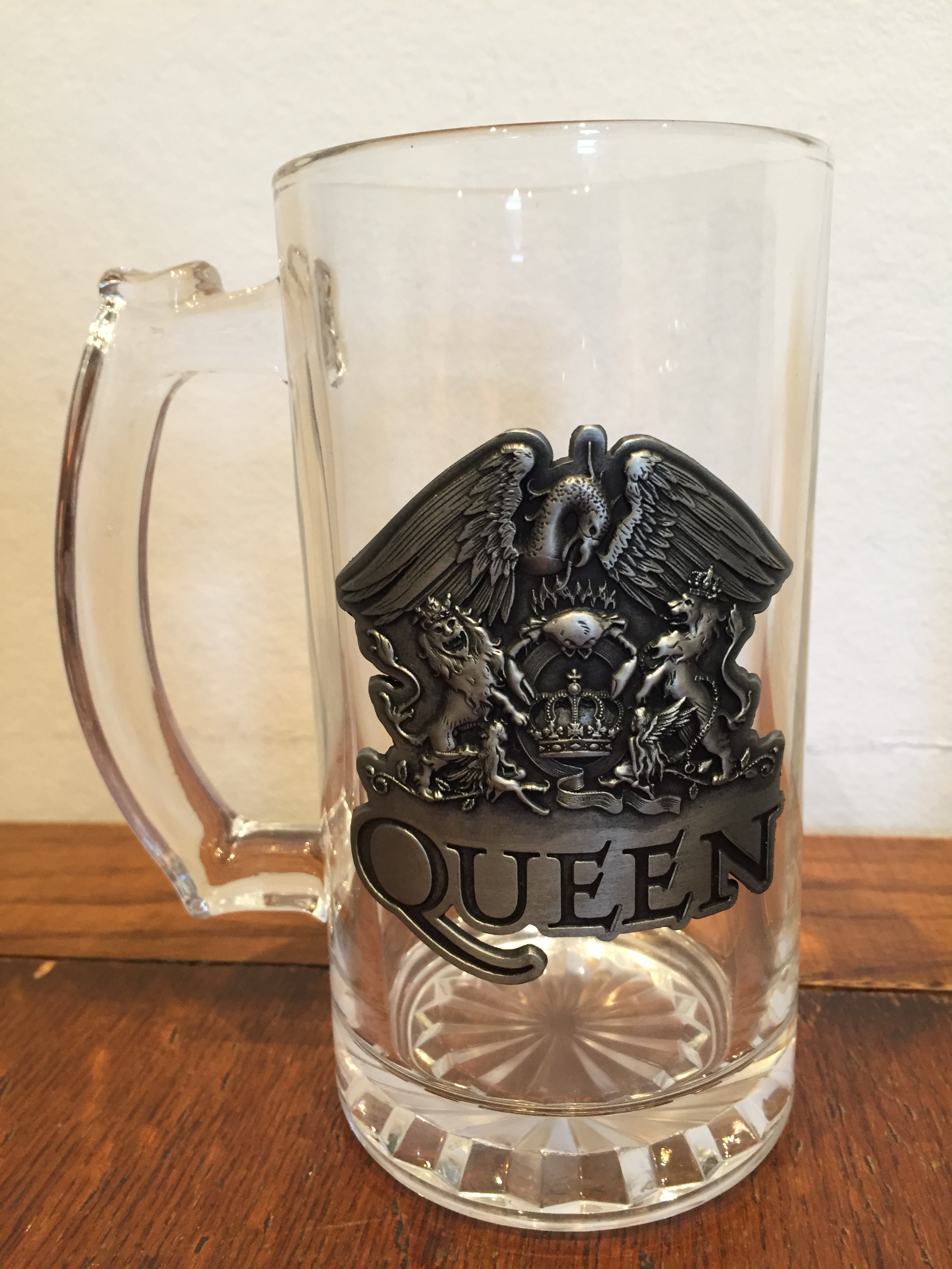 A large glass stein, basically a pint glass with a handle. On the side is a large metal version of the Queen crest. The crest has a large phoenix sitting on top of a large letter Q with a crown in the middle of it. 2 lions, one on each side, are holding up the letter Q with their front legs while standing on their back legs. The band's name is below the crest.