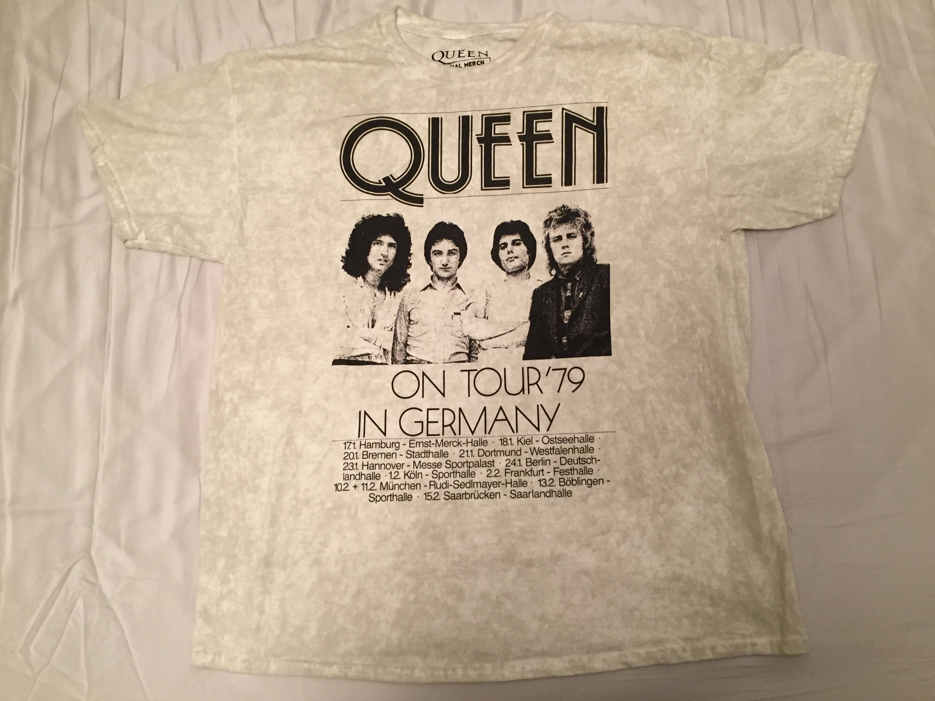 Short-sleeved t-shirt in a dirty white colour. From top to bottom, the black coloured design has the band name Queen in large letters, then a photo of Brian, John, Freddie and Roger, then large capital letters saying On Tour 79 In Germany, and finally 6 lines of much smaller text with the dates and venues from the tour.