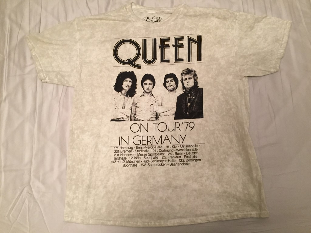 Short-sleeved t-shirt in a dirty white colour. From top to bottom, the black coloured design has the band name Queen in large letters, then a photo of Brian, John, Freddie and Roger, then large capital letters saying On Tour 79 In Germany, and finally 6 lines of much smaller text with the dates and venues from the tour.