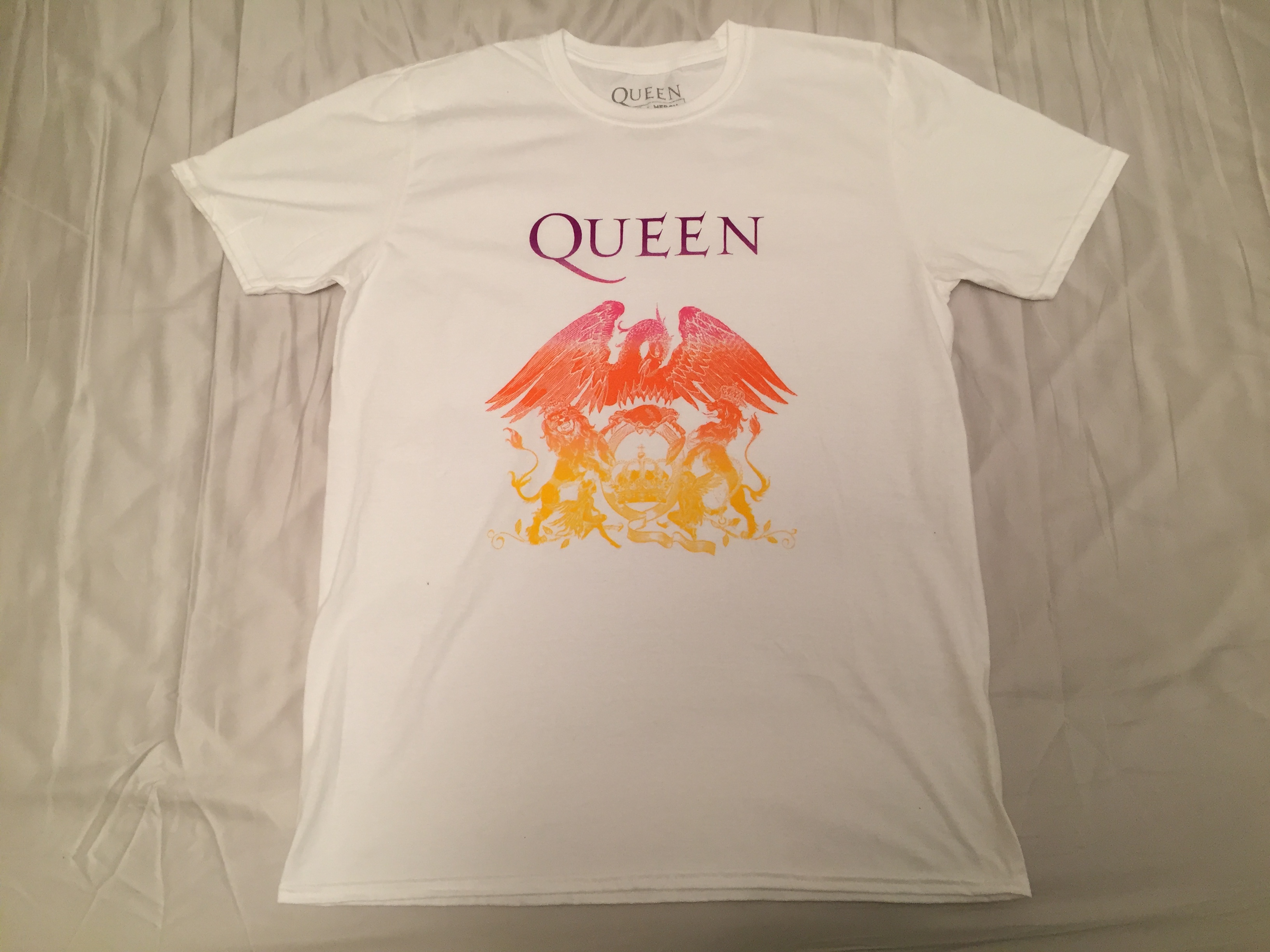 Short sleeved white t-shirt with the crest of the band Queen in the centre, which flows through pink, red, orange and yellow colours from top to bottom. At the top is the band's name. Below this, a large phoenix spreads its wings over the top of a crab, which is sitting on a letter Q with a crown in the middle of it. 2 lions, one on each side, are holding up the letter Q with their front legs while standing on their back legs. 2 fairies are seated either side of the Q at the bottom.