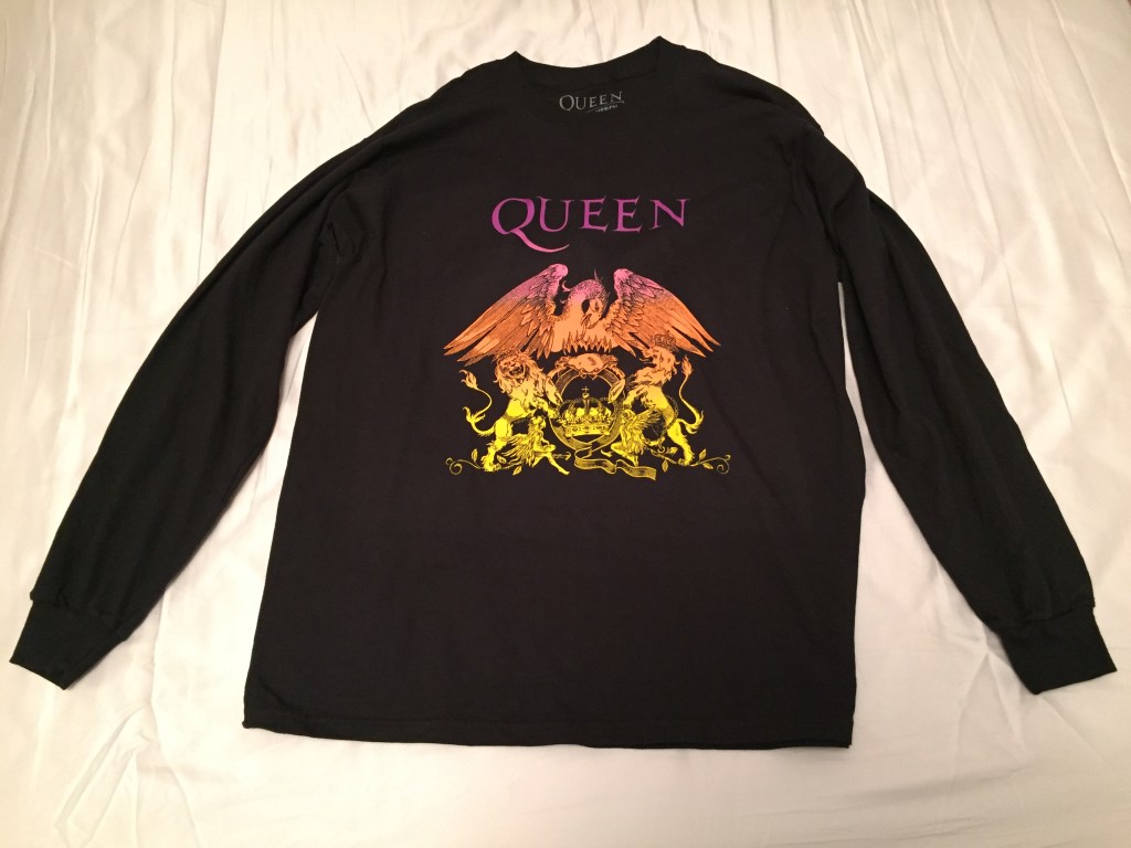 Long sleeved black t-shirt with the crest of the band Queen in the centre, which flows through pink, red, orange and yellow colours from top to bottom. At the top is the band's name. Below this, a large phoenix spreads its wings over the top of a crab, which is sitting on a letter Q with a crown in the middle of it. 2 lions, one on each side, are holding up the letter Q with their front legs while standing on their back legs. 2 fairies are seated either side of the Q at the bottom.