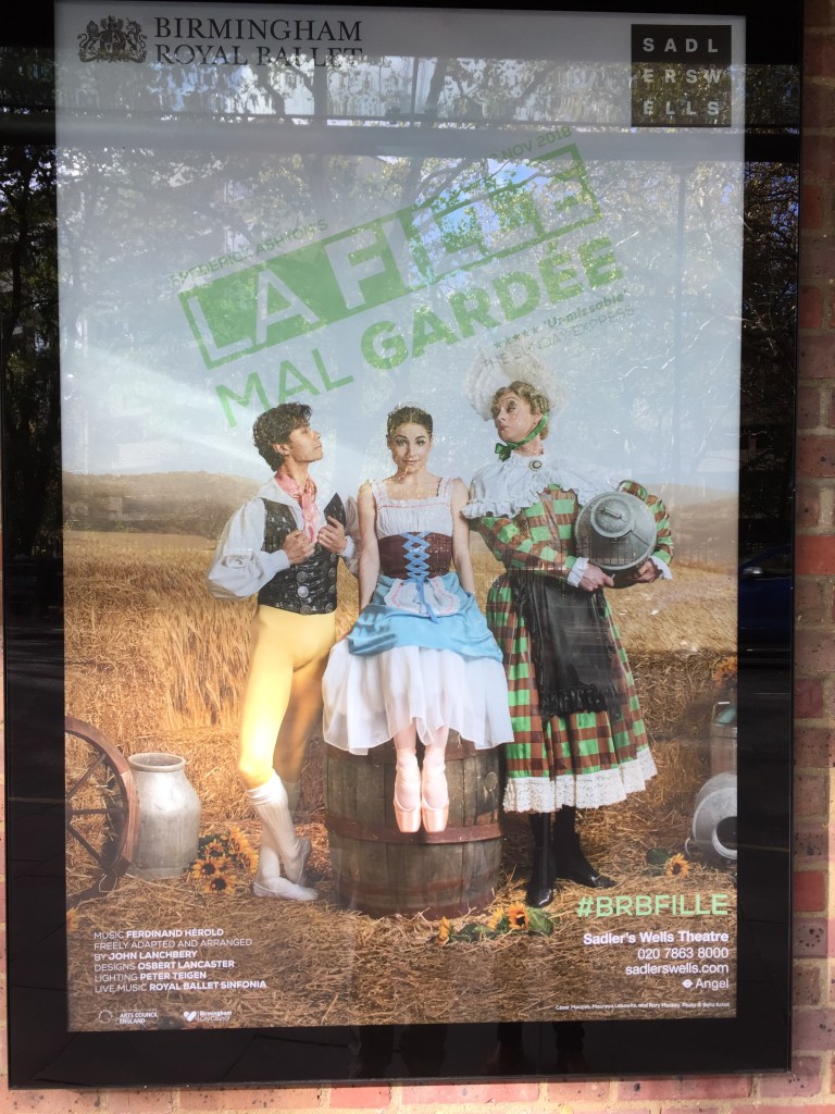 Poster for La Fille Mal Gardée, showing the 3 central characters in a corn field. The handsome man on the left has tight yellow trousers, a black jacket over a shirt with white sleeves, and a pink ribbon tied around his neck. He is looking lovingly at the young lady sitting on a barrel in the centre, whose dress has a flowing white skirt with a blue layer of fabric on top and a black chest section with criss-crossing blue fabric up the centre. To her right, her stern looking mother is wearing a large green and red chequered dress with decorative white edging at the hem and shoulders, and is holding a grey hat.