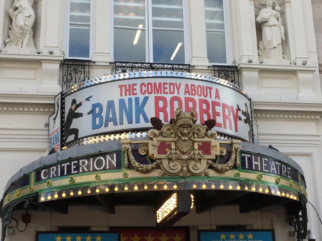 Circular canopy above the entrance to the Criterion Theatre. A small grinning face forms the centrepiece of the ornate gold crest in the middle, either side of which are the words Criterion and Theatre in white letters on a black background. Above this, on another curved panel, is the banner for Comedy About A Bank Robbery. The title of the play is in red and blue capital lettering, with a person dressed in black on each side reaching out with gloved hands to try and steal a letter from the text.